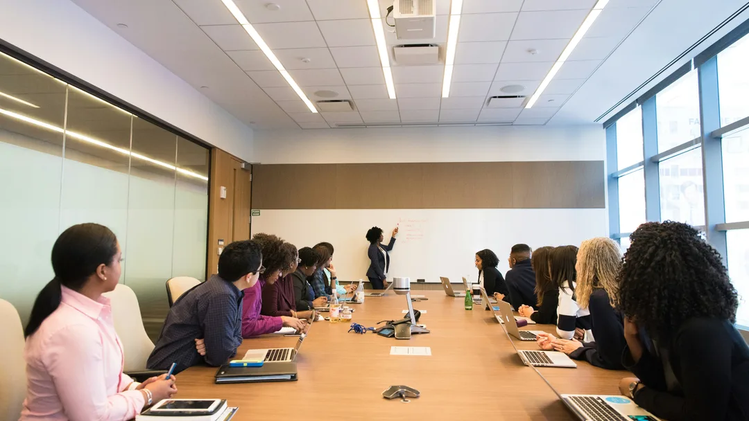 Bright meeting room with people seated with laptops along a long wooden table, facing a woman standing at a whiteboard at the far end