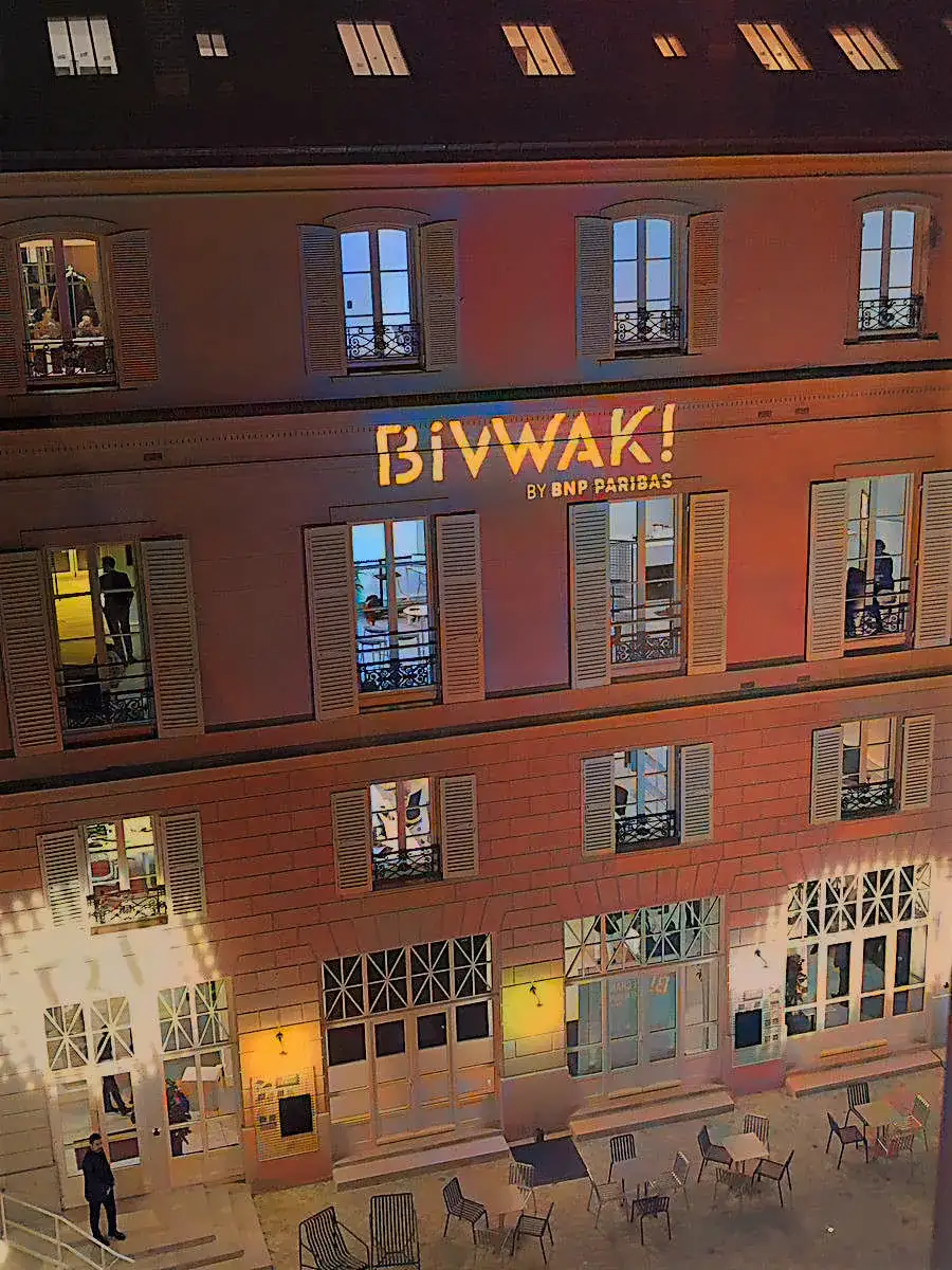 La façade de Bivwak! by BNP Paribas à Paris : son nom au mur parmi les fenêtres éclairées, une terrasse de chaises en contrebas